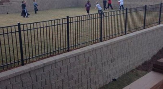 People walking on a grassy slope beside a black metal fence and stone retaining wall.