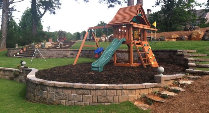 Backyard playground with wooden playset, slide, and circular stone retaining wall in a grassy yard