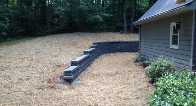 Retaining wall of stacked gray blocks beside a house, bordering a dry yard and wooded background
