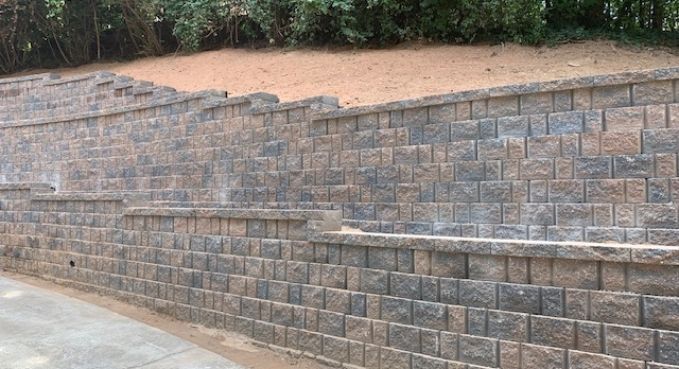 Stone retaining wall with steps along a sloped roadside beside greenery