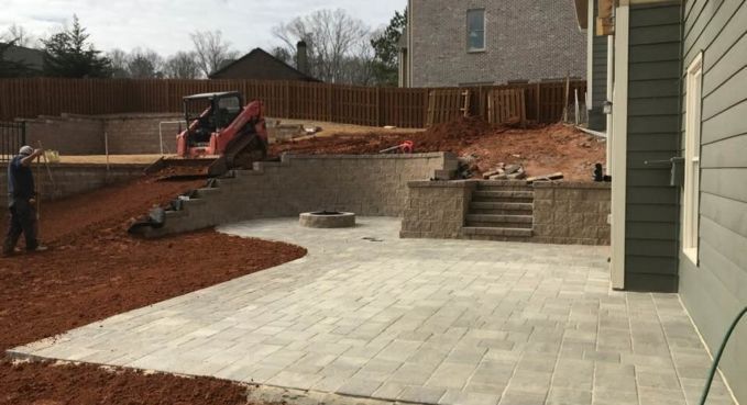 Backyard patio under construction with excavator, retaining wall, pavers, and red mulch