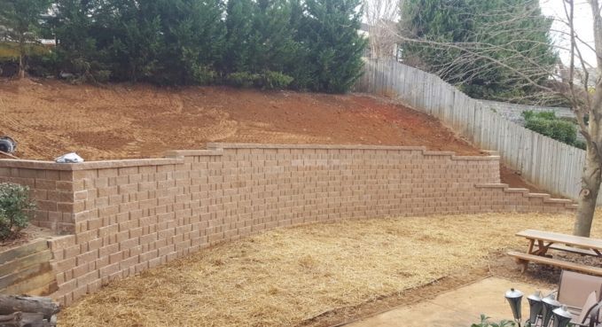 Backyard with a newly built tan retaining wall below a sloped, dirt-covered hill and trees above