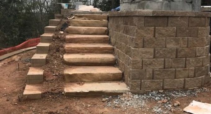 Unfinished outdoor stone steps beside a retaining wall on a dirt slope with scattered debris.