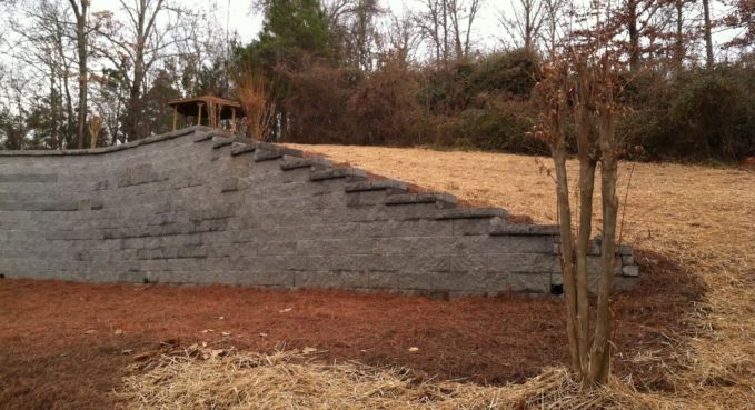 Stone retaining wall on a sloped, wooded hillside with bare trees and brown ground cover