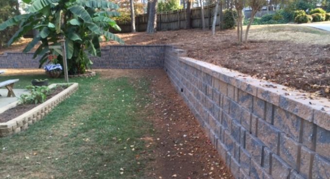 Sloped grassy yard beside a brick retaining wall and dirt embankment, with a tree on the left.