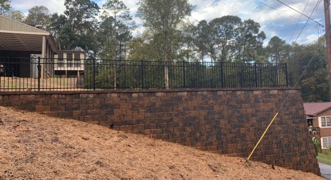 Brick retaining wall on a sloped yard beside a house, with trees and a fenced deck above.