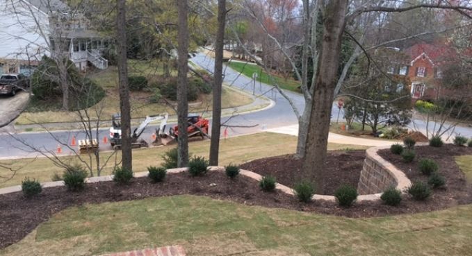 Driveway work in progress on a curving road, with two utility trucks and two large trees in a landscaped front yard.