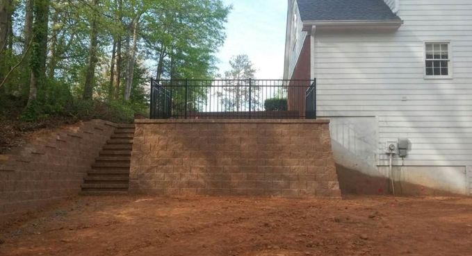 Brick retaining wall with stairs beside a white house and wooded backyard area