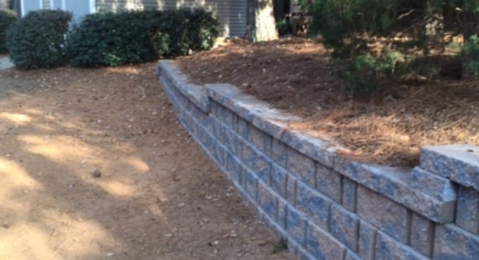 Curved gray stone retaining wall along a dirt path beside shrubs and mulch in a landscaped yard