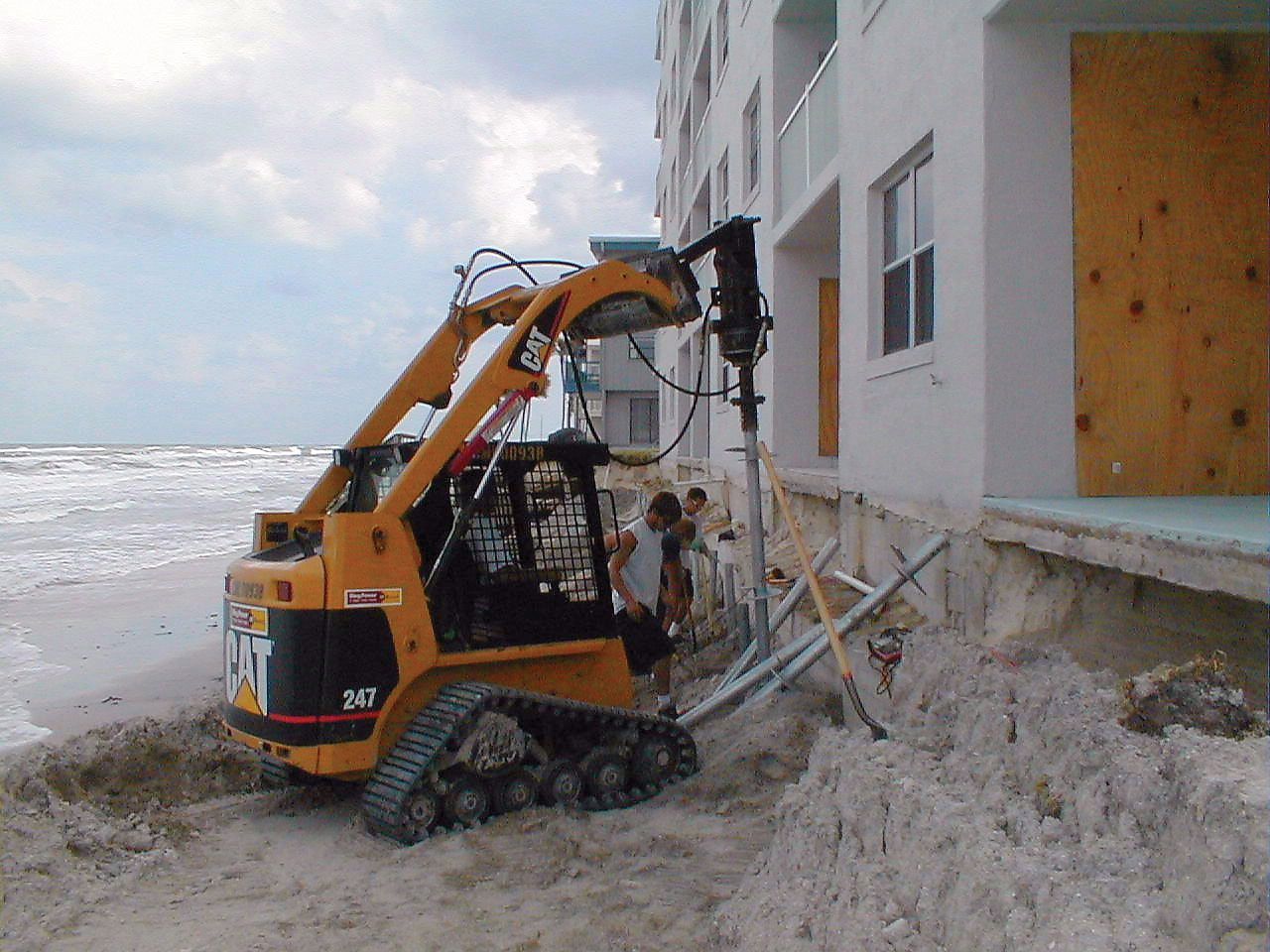Orange skid-steer loader working beside a beach house, digging at a sand-covered foundation