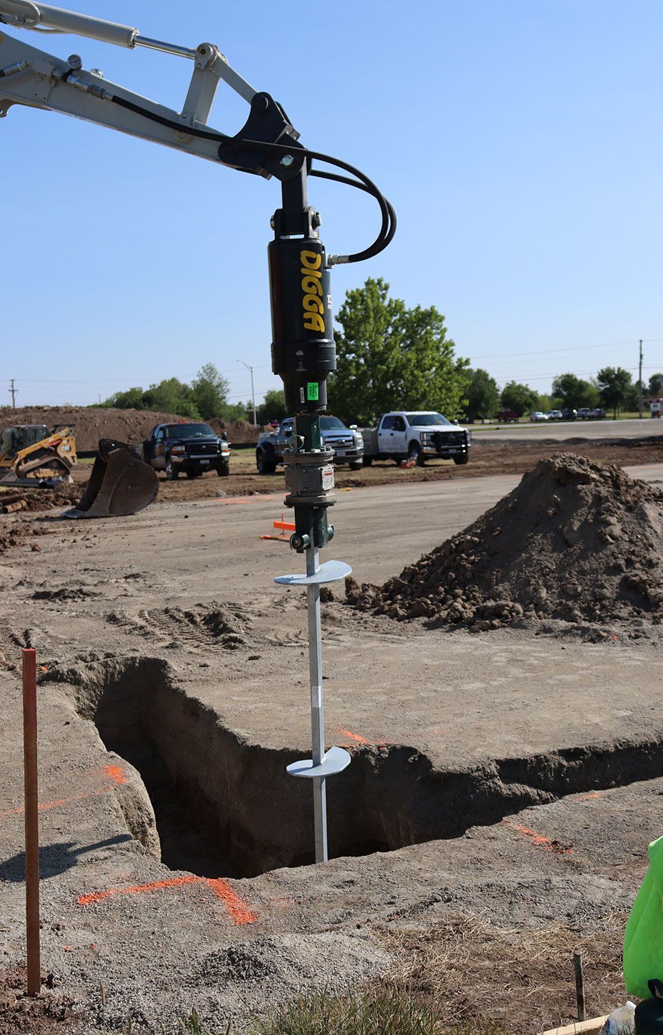 Construction auger drilling a post hole beside an excavator at a dirt site