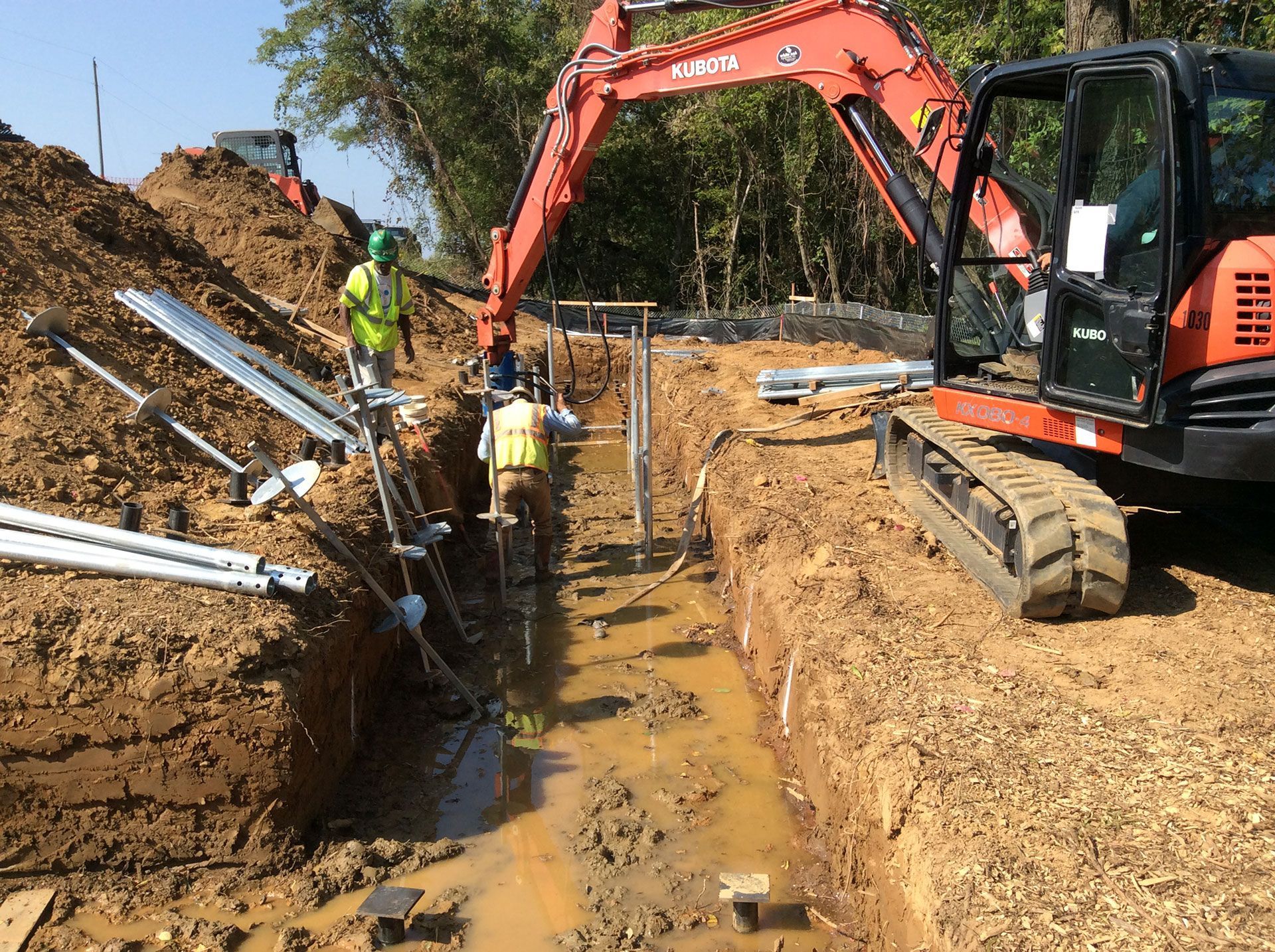Construction crew working in a muddy trench beside an orange excavator and metal pipes