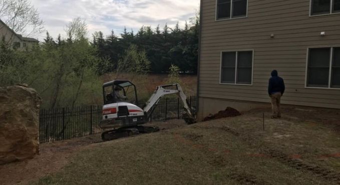 Mini excavator and dirt beside a house, with a person walking nearby on a sloped yard.