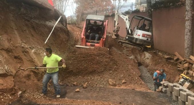 Workers clearing a large dirt landslide on a steep road with a backhoe and truck