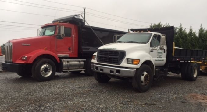 Red dump truck and white flatbed truck parked side by side on a gravel lot