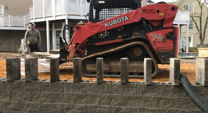 Red Kubota skid steer on a construction site beside concrete footings and a worker in the background.