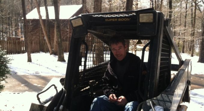 Person sitting in a black snowplow or utility vehicle in a snowy yard, facing the camera