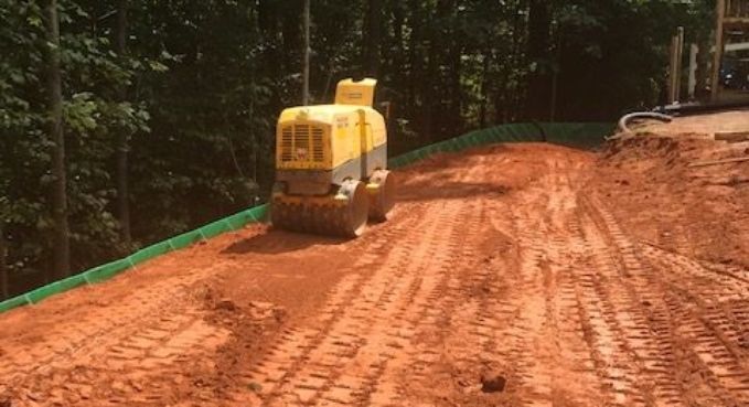 Small yellow construction roller compacting a red dirt road on a forested hillside