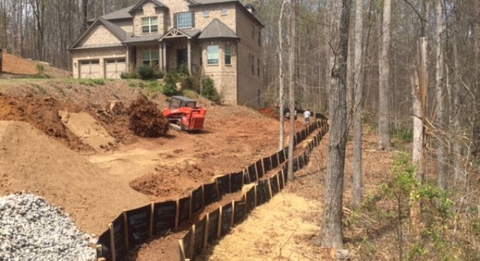 House on a steep wooded lot with excavation work, an orange skid steer, and a black erosion-control barrier.