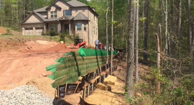 House under construction beside a wooded lot, with workers, green silt fence, and dirt slope.