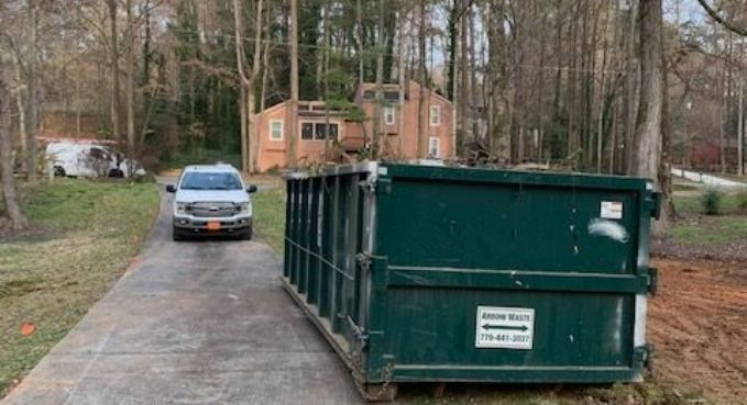 Green dumpster on a driveway beside a car, with a wooded house lot in the background.