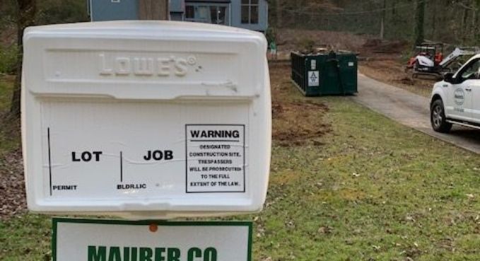 White Lowe's dumpster on grass with lot/job labels, near a green dumpster and parked vehicles.