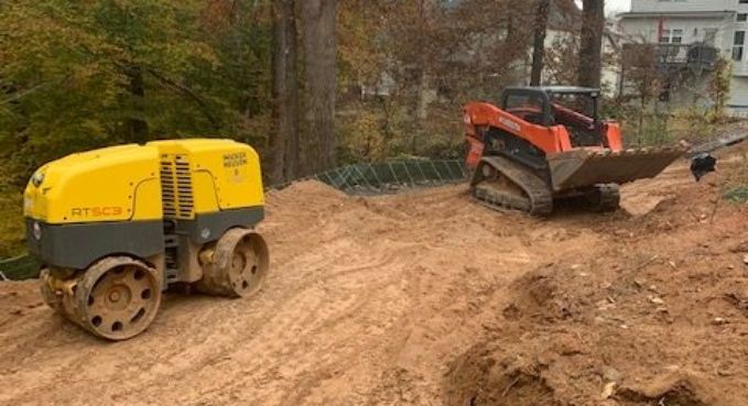 Yellow road roller and orange skid steer on a muddy construction site surrounded by trees.