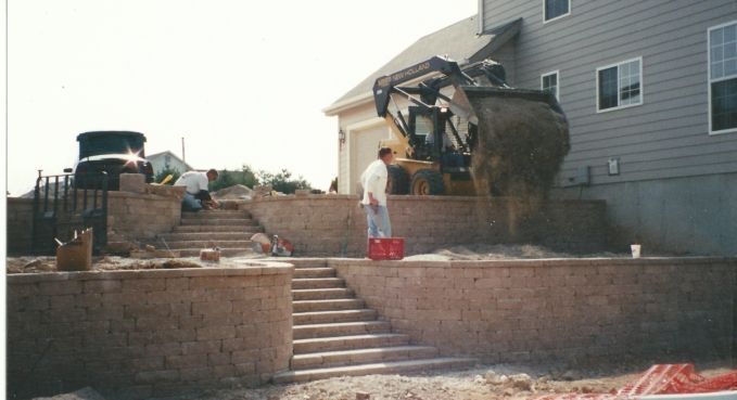 Construction crew using a skid steer to clear dirt beside a house and brick retaining wall