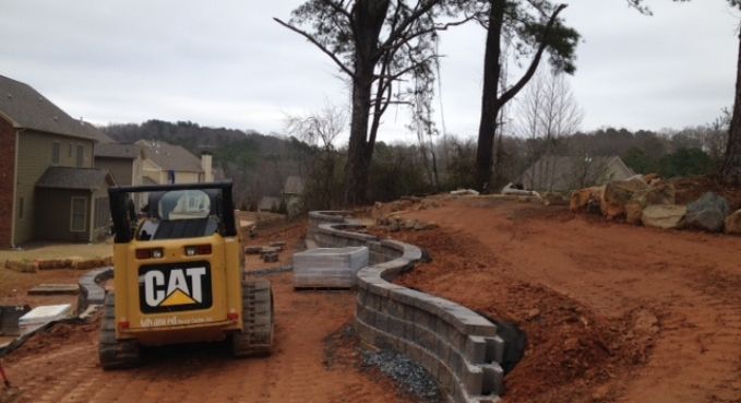 CAT excavator installing curved retaining wall along a red dirt driveway beside a house