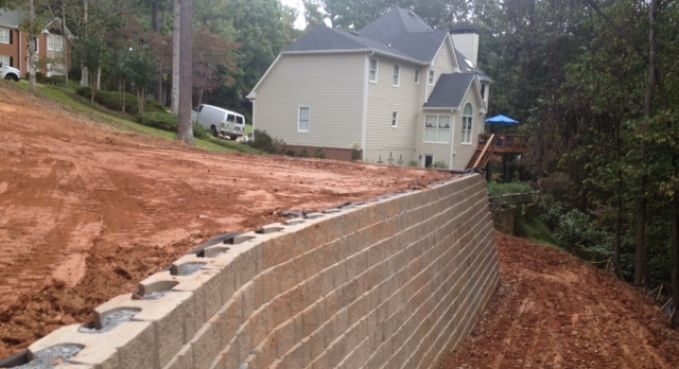 Retaining wall beside a sloped dirt lot behind a house, with a wooded area in the background