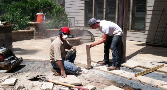 Two workers laying paving stones in a backyard patio area beside a house