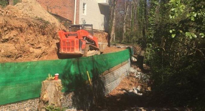 Orange excavator on a dirt hillside beside a green retaining wall in a wooded area