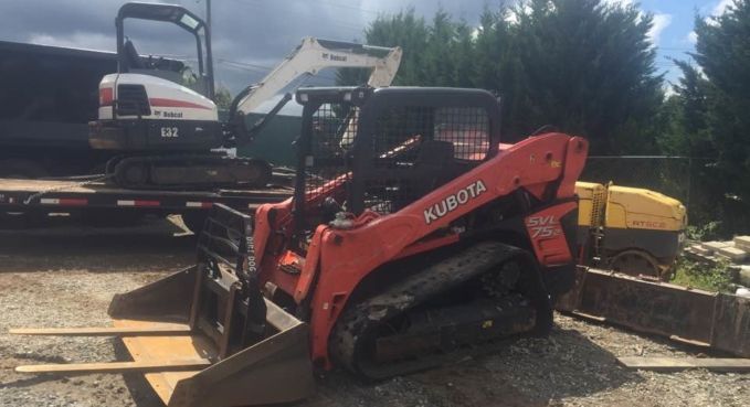 Red Kubota mini excavator parked on a flatbed trailer in a gravel yard.