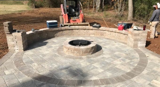 Circular stone patio with a central fire pit under construction, with a mini excavator and workers nearby