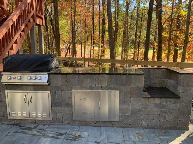 Outdoor stone patio with built-in grill, stainless cabinets, and a seating ledge by a wooded deck stairway