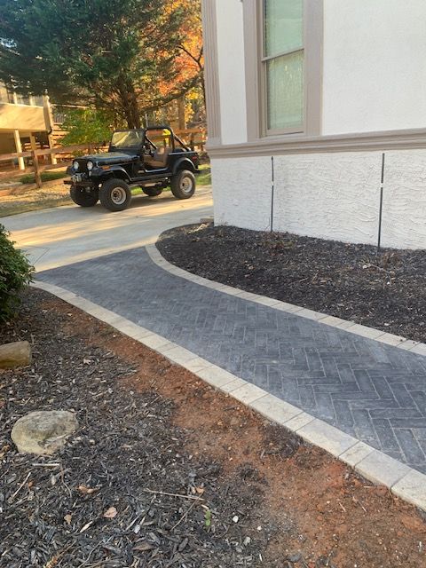 Outdoor walkway beside a building with a paver path, mulch beds, and a utility vehicle in the background