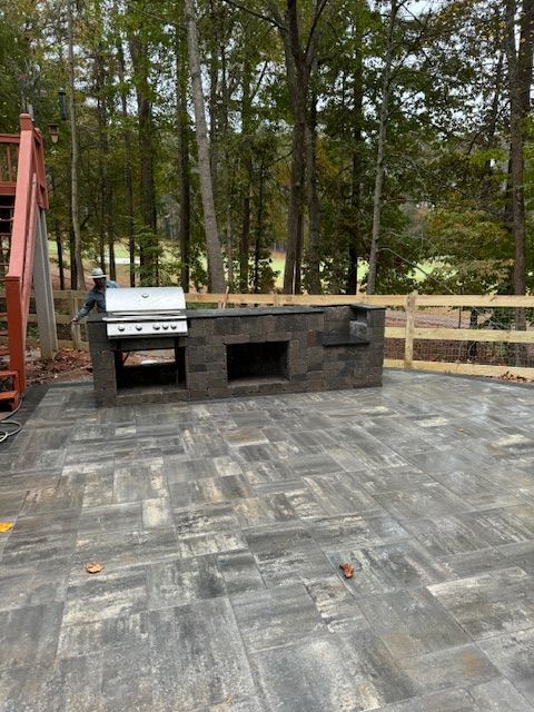 Outdoor patio with gray pavers, built-in grill and stone counter, surrounded by a wooden fence and trees.