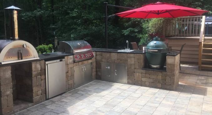 Outdoor patio with brick pizza oven, gas grill, green smoker, and red umbrella on a stone terrace