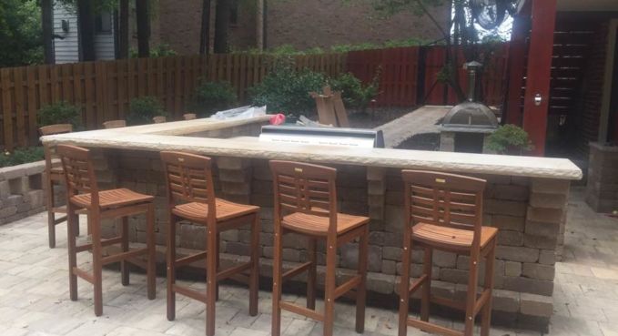 Outdoor stone patio bar with four wooden stools and a light-colored countertop under trees.