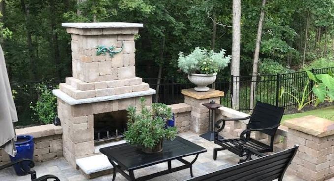 Backyard patio with stone fireplace, black chairs, and potted plants on a deck overlooking trees