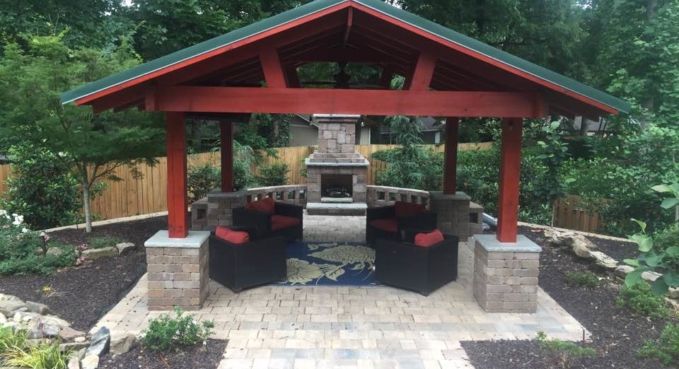 Covered outdoor seating area with red roof, stone pillars, and black chairs in a landscaped garden