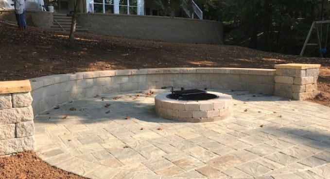 Stone patio with built-in circular fire pit and curved retaining wall in a wooded backyard