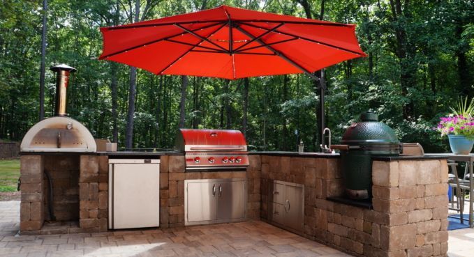 Outdoor brick grill station with red umbrella, pizza oven, gas grill, sink, and counter in a wooded yard