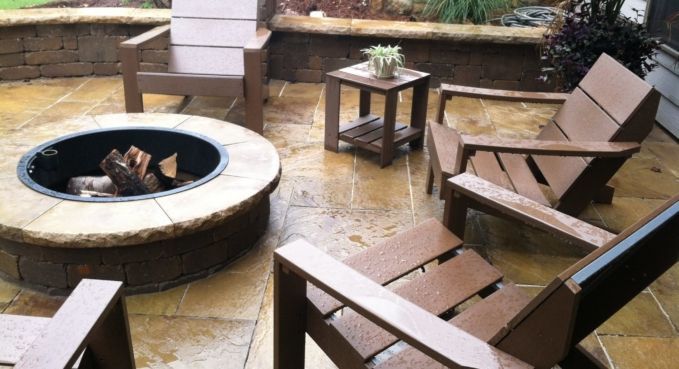 Patio with a round fire pit, wooden lounge chairs, and a small side table on wet stone paving