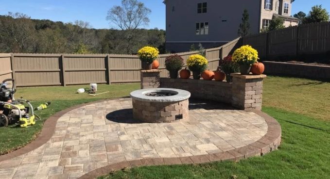 Circular stone fire pit on a curved patio in a fenced backyard with flower pots and lawn