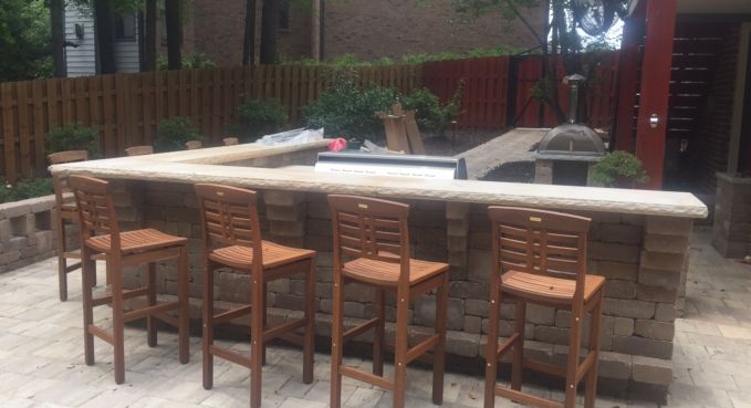 Outdoor stone patio bar with four wooden stools and a built-in counter in a backyard garden area