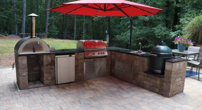 Outdoor brick patio with a wood-fired pizza oven, grill, sink, and red umbrella in a backyard setting