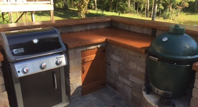 Outdoor patio with a stainless grill and green ceramic smoker beside a brick counter and wooden bench seat