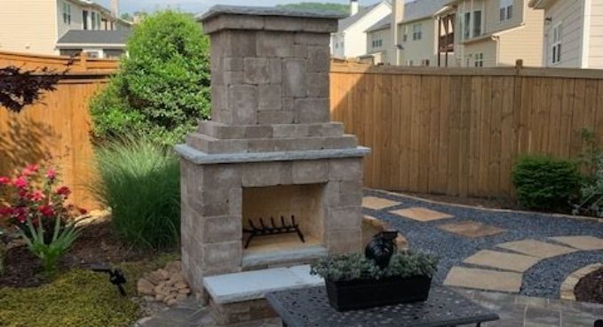 Stone outdoor fireplace in a landscaped backyard patio with pavers, plants, and a wooden fence.