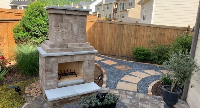 Stone outdoor fireplace beside a curved paver patio and wooden fence in a backyard garden